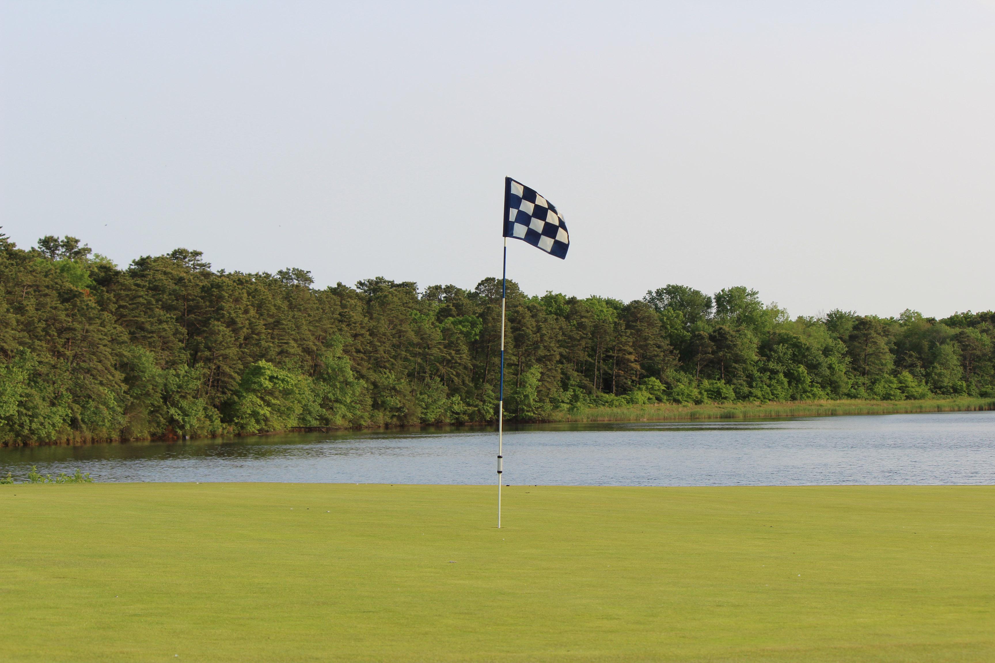 10th Green at Ocean Acres Country Club. In the foreground, is a blue and white checkered flag. Beyond the green is Holiday Lake that is bordered by large and dense trees, mostly pine trees.
