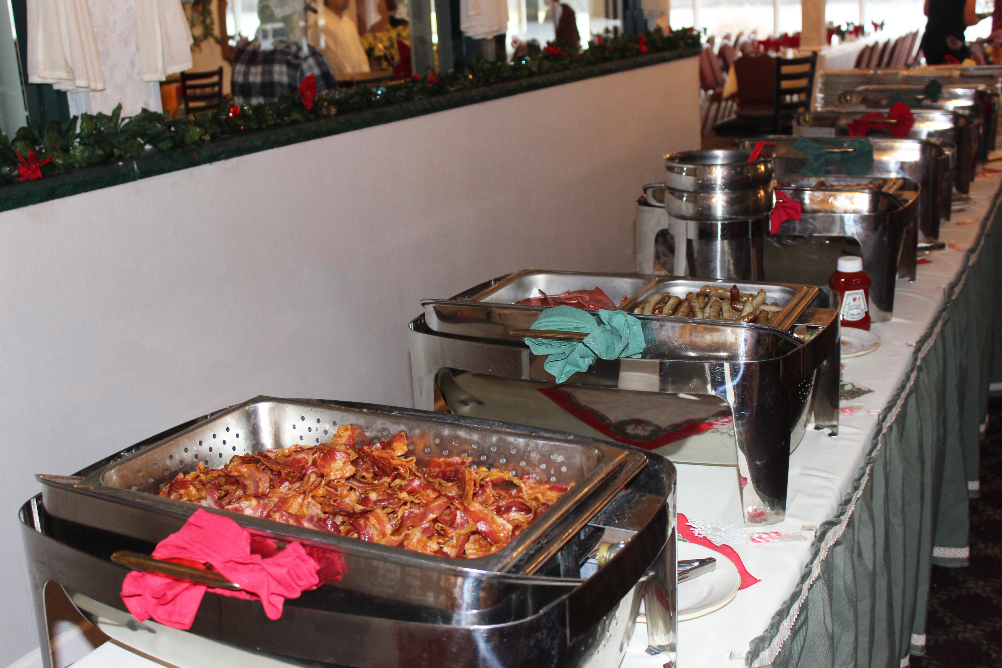 Along a white wall, there are eight metal food chafers set on a skirted table and decorated with a holiday theme of red and green. Bacon, sausage links, and pork roll can be seen in the first chafers.