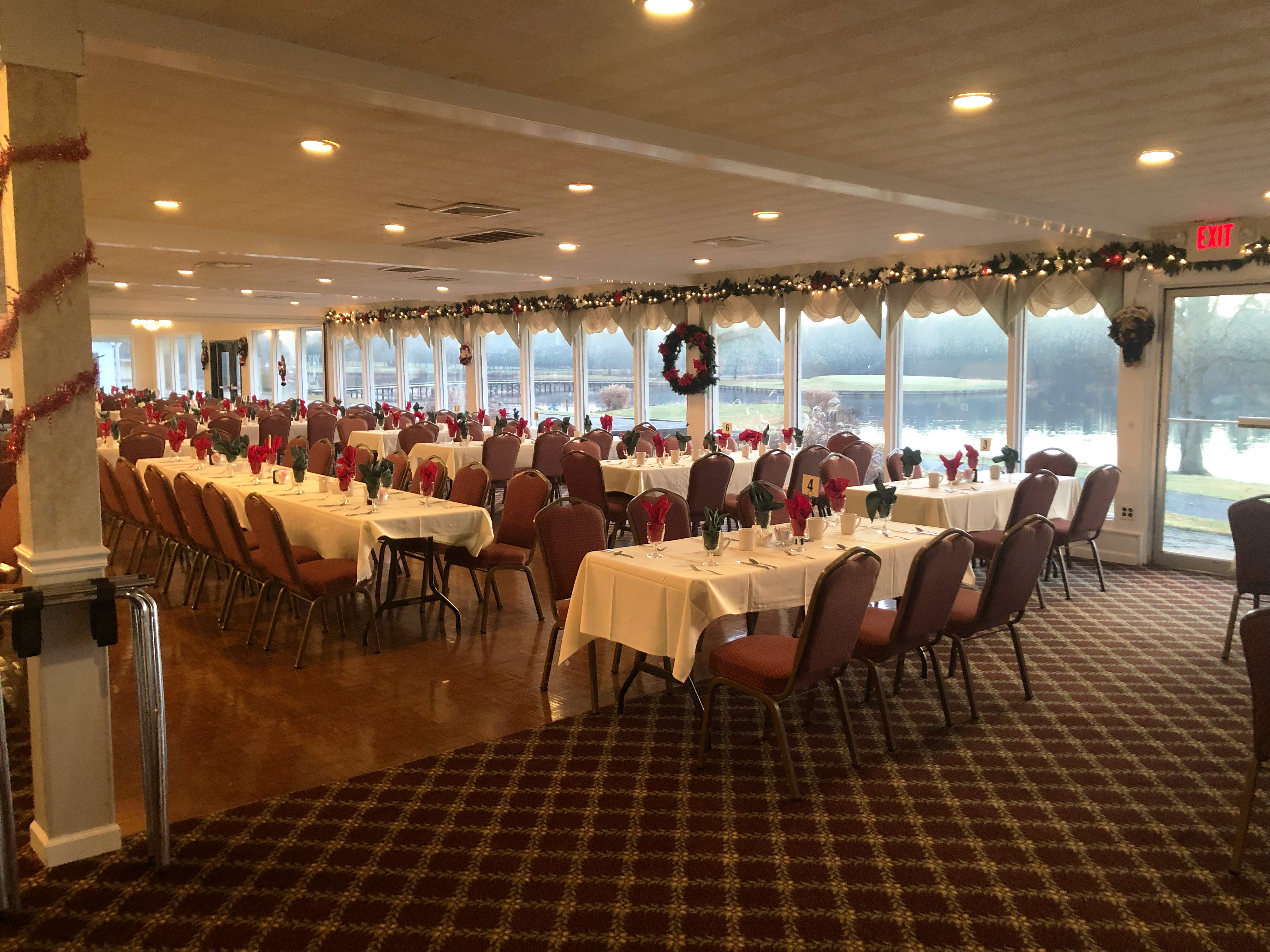 A banquet room set with various rectangular tables set with white table cloths, drinking glasses, and red and green napkins fill the foreground. The room is also decorated with a Christmas wreath, and garland lit with Christmas lights. Through the windows, the morning light shines on a golf course, particularly a green that is positioned across a lake.