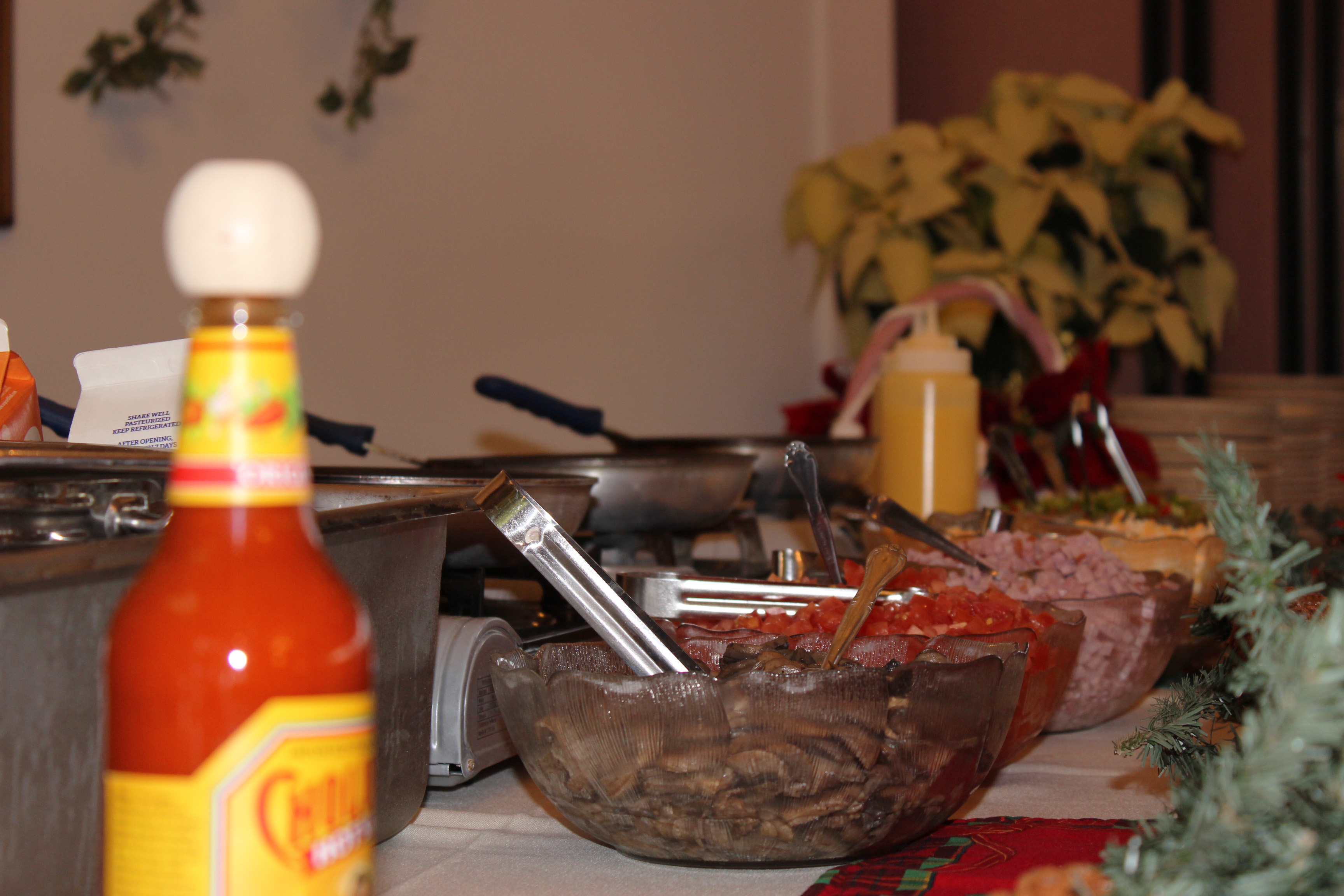 Cholula Hot Sauce and big bowls of sliced mushrooms, diced tomatoes and ham, shredded cheese, and diced red and green peppers fill the foreground. A metal insulated container for eggs and three frying pans can also be seen. The table is decorated with garland and flowers.