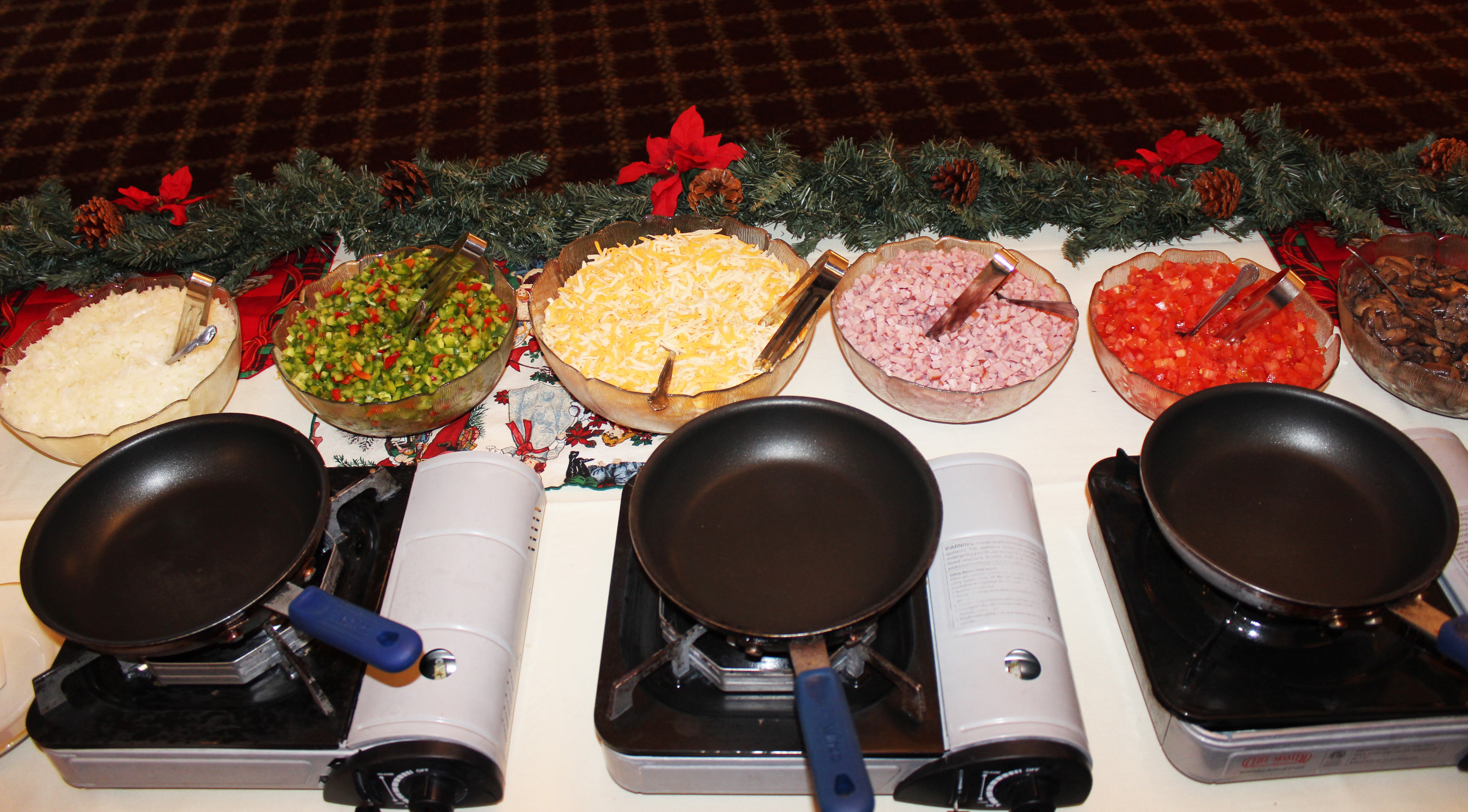 Above view of three frying pans in front of big bowls of diced onions, diced red and green peppers, shredded cheese, diced ham, diced tomatoes, and sliced mushrooms. Garland decorated with pine cones and red ribbons border the table.