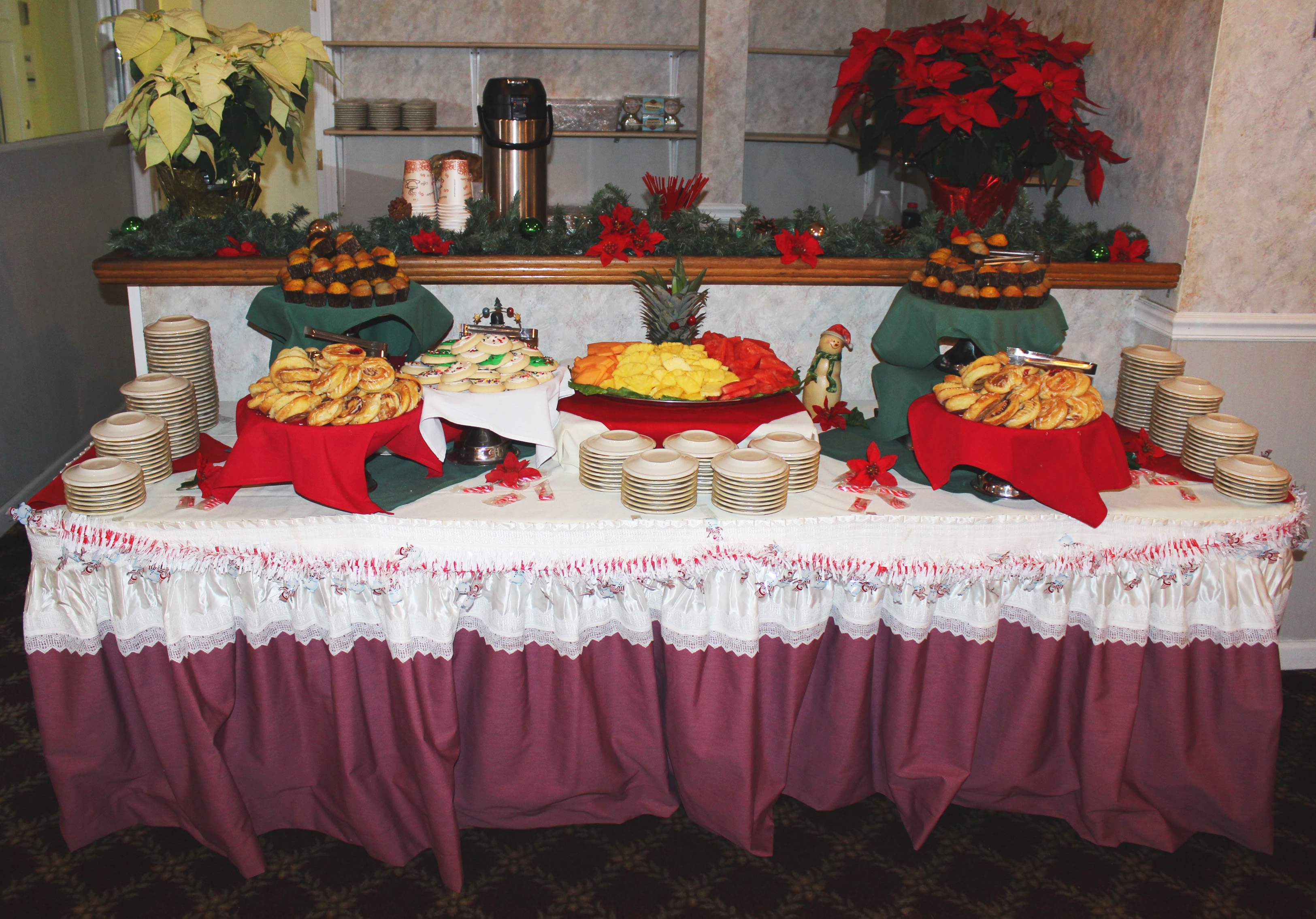Upon a red-skirted and holiday-decorated table, are two trays of assorted muffins, two trays of assorted danish, a tray of cookies, and a display of fresh fruit including pineapple, cantaloupe, and watermelon. Behind the table, is a bar, decorated with garland.