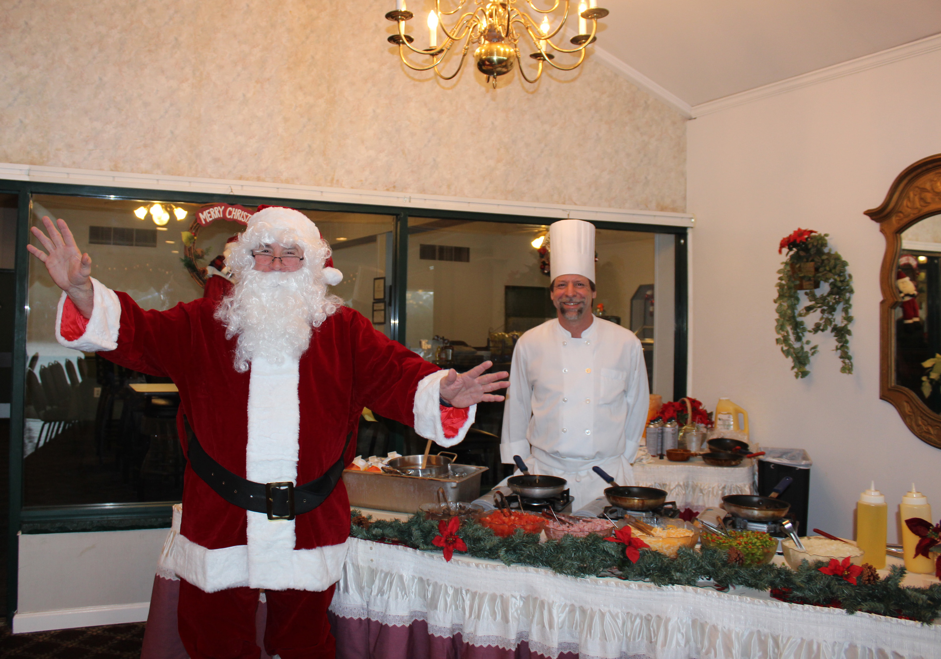 Santa Claus stands in front of a table upon which there are three frying pans, along with cooking supplies, eggs, and big bowls of mushrooms, tomatoes, cheese, red and green peppers, and onions. Behind the table stands a chef smiling and wearing chef's whites and a chef toque. Christmas decorations can be seen as well.