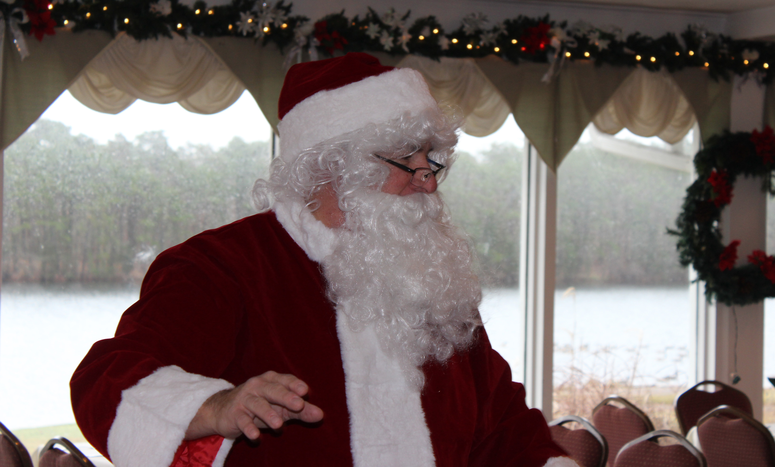 Santa Claus in his big red suit is looking to the right. In the background, there are a number of paneled windows through which a lake and pine trees are visible.