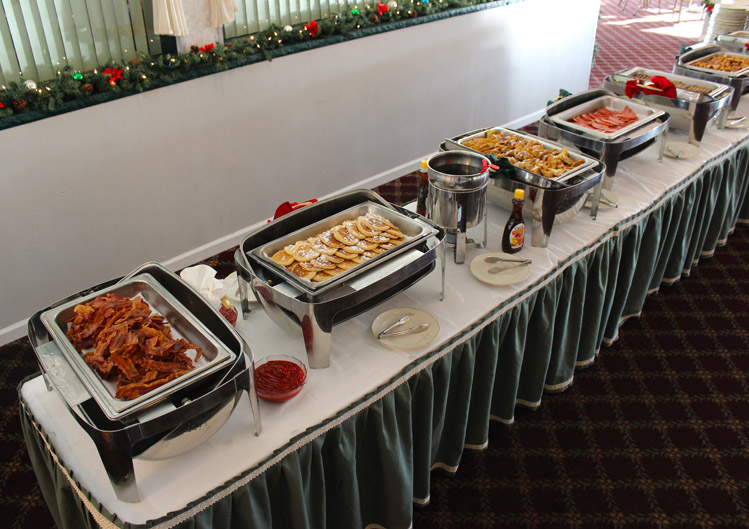 Six chafers lined up on a banquet table with Christmas garland and decorations. The chafers are filled with various breakfast items including bacon, pancakes, french toast, pork roll, and breakfast potatoes. Syrup and other condiments are also on the table.