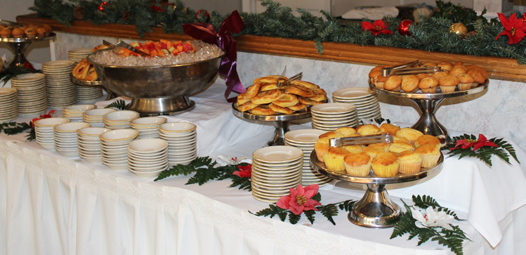 A table adorned with Christmas ornaments, garland and poinsettias, holds several platters of a variety of pastries and fruits.