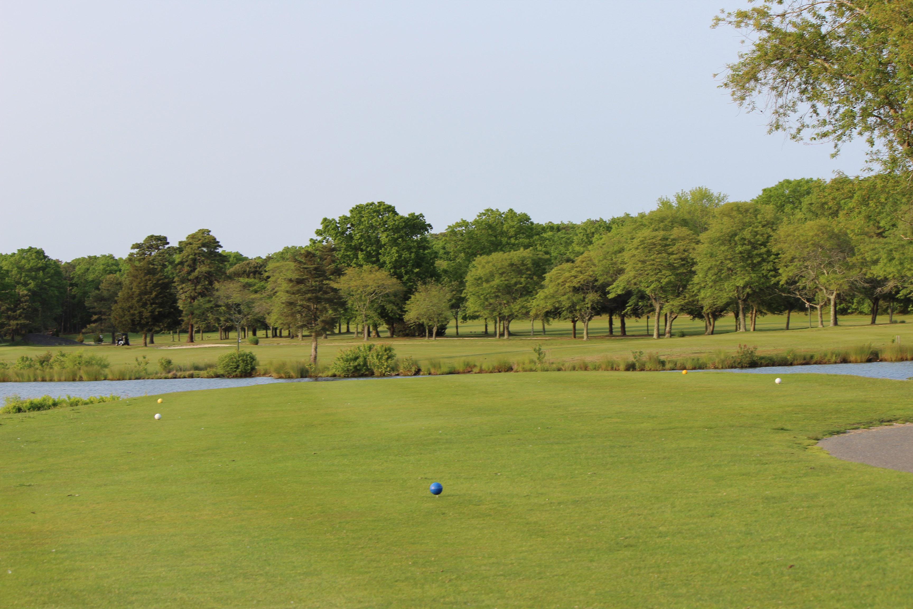 The view from the tee box on Hole No. 11 at Ocean Acres Country Club. Blue, white, and yellow tee markers are positioned on the tee box. Where the tee box ends, Holiday Lake begins. The fairway is on the other side of the lake. The treeline borders the right side of the hole.