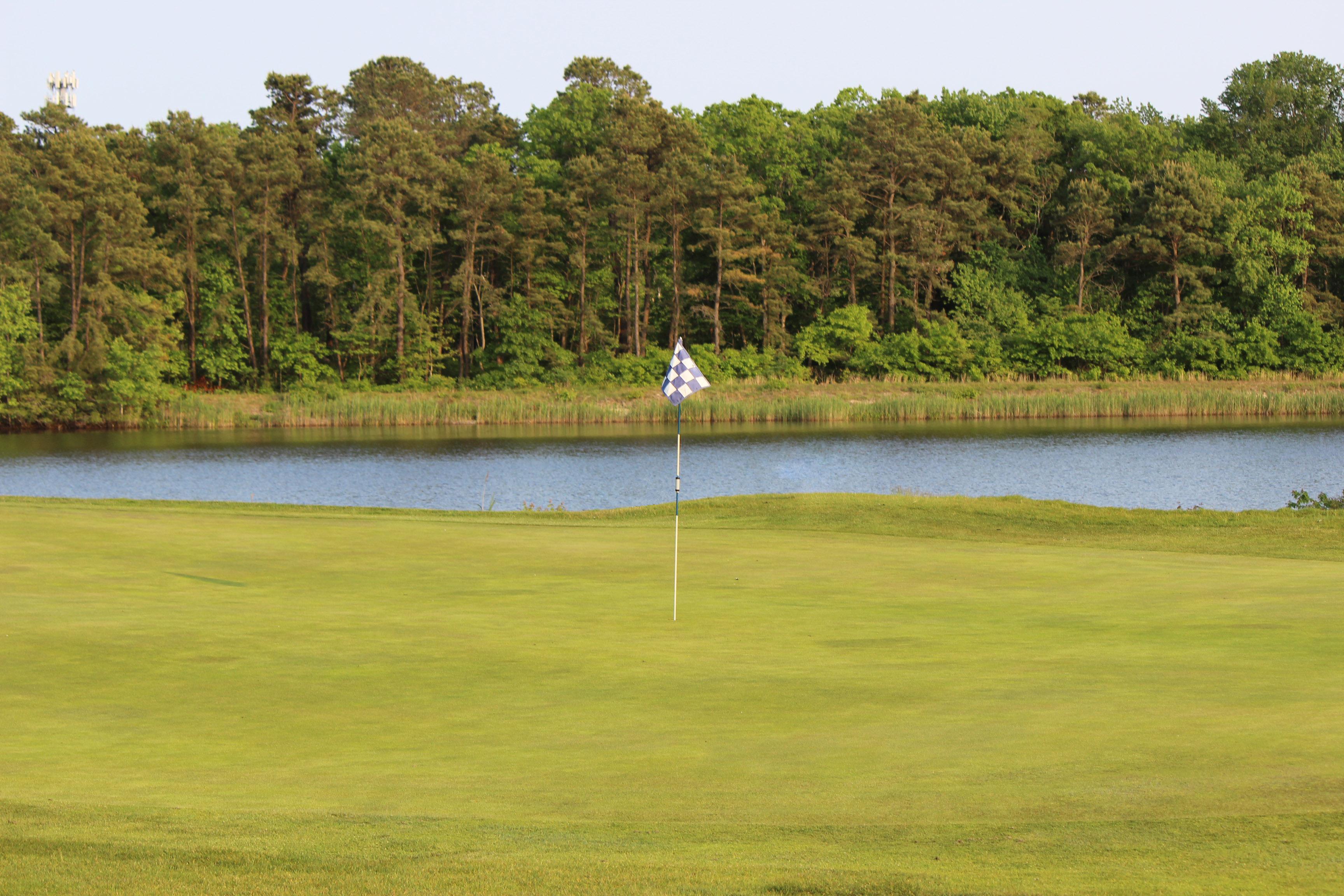 The green on Hole No. 9 at Ocean Acres Country Club. The flag stick is centered on the green and the flag is checkered white and blue. Rough and fringe surround the green. Beyond the green is Holiday Lake which is bordered by reeds and dense pine trees.