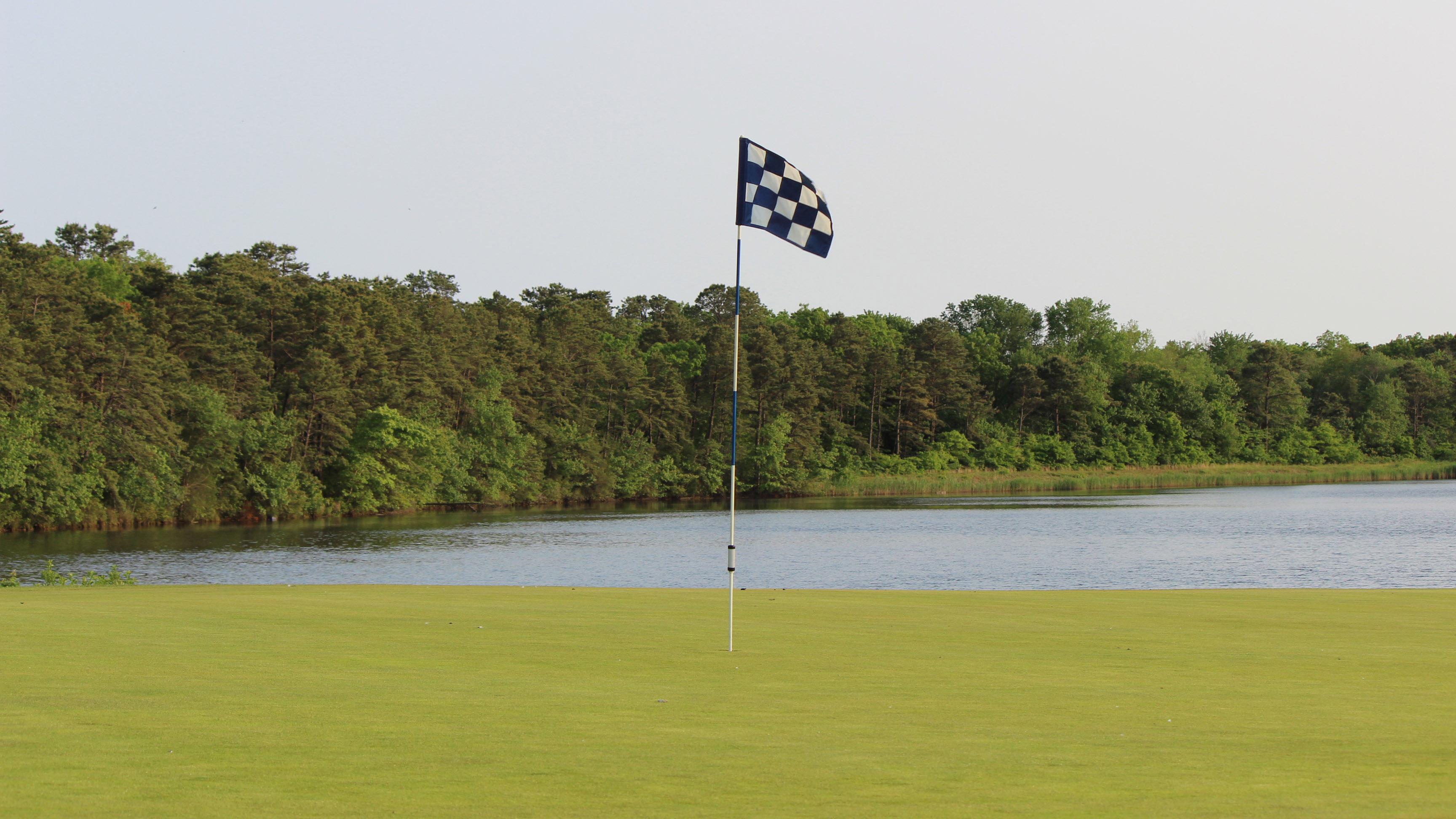 10th Green at Ocean Acres Country Club. In the foreground, is a blue and white checkered flag. Beyond the green is Holiday Lake that is bordered by large and dense trees, mostly pine trees.