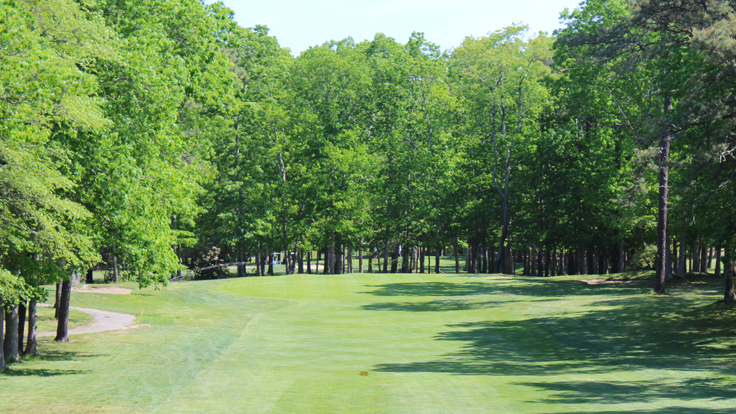 The approach to the 12th hole at Ocean Acres Country Club. Several trees outline the fairway down towards the green with more trees around the green casting shadows upon it. There are sunny and bright skies above.