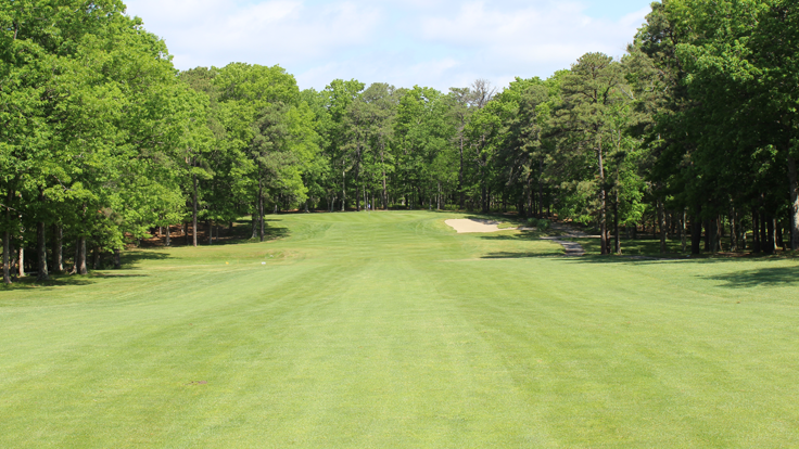 The approach down the fairway to the par 5 15th hole at Ocean Acres Country Club. The fairway slopes down before sloping back upwards 30 yards from the green. The fairway is lined on both sides by dense trees and a right greenside bunker protects the green.