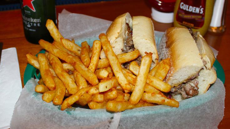 Cheesesteak in a steak roll cut in half and served with a good helping of french fries. A bottle of Heineken and Ketchup and Mustard bottles can be seen behind the food.
