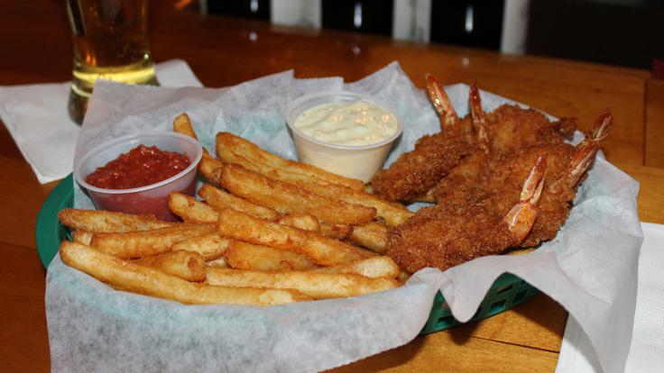 5 fried jumbo shrimp served in a green food basket with a plain liner. French fries, marinara sauce, and tarter sauce are also in the basket.