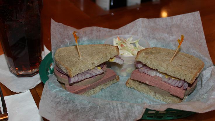 Liverwurst sandwich with red onions and mustard on rye bread. The sandwich is cut in half with frill picks holding together each half. The sandwich is served with coleslaw.