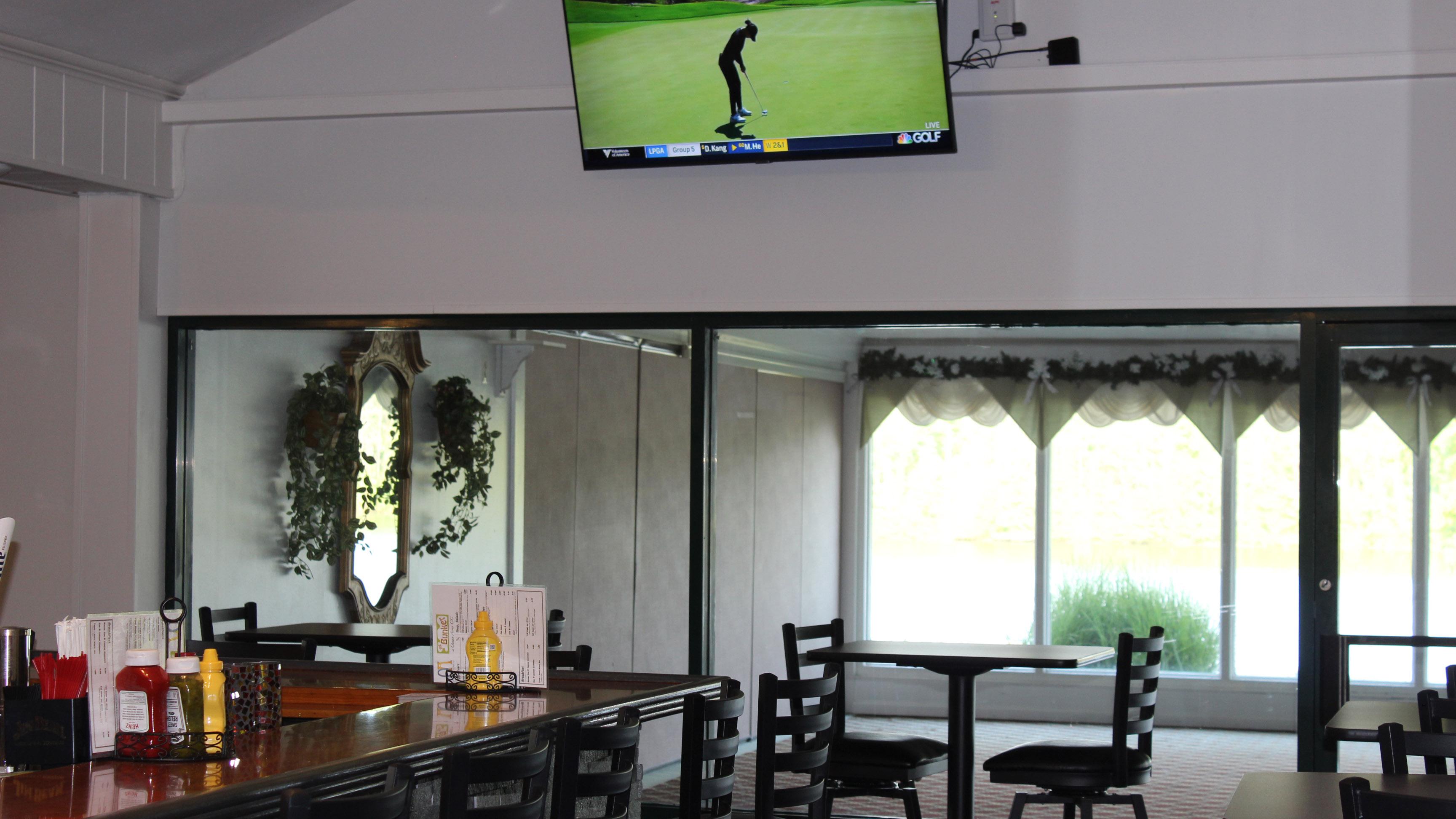 The bar counter can be seen in the left side of the picture. The focus is on the television hanging on the wall and turned on to the Golf Channel. In the background, there are high top tables and the windows look out to the golf course.