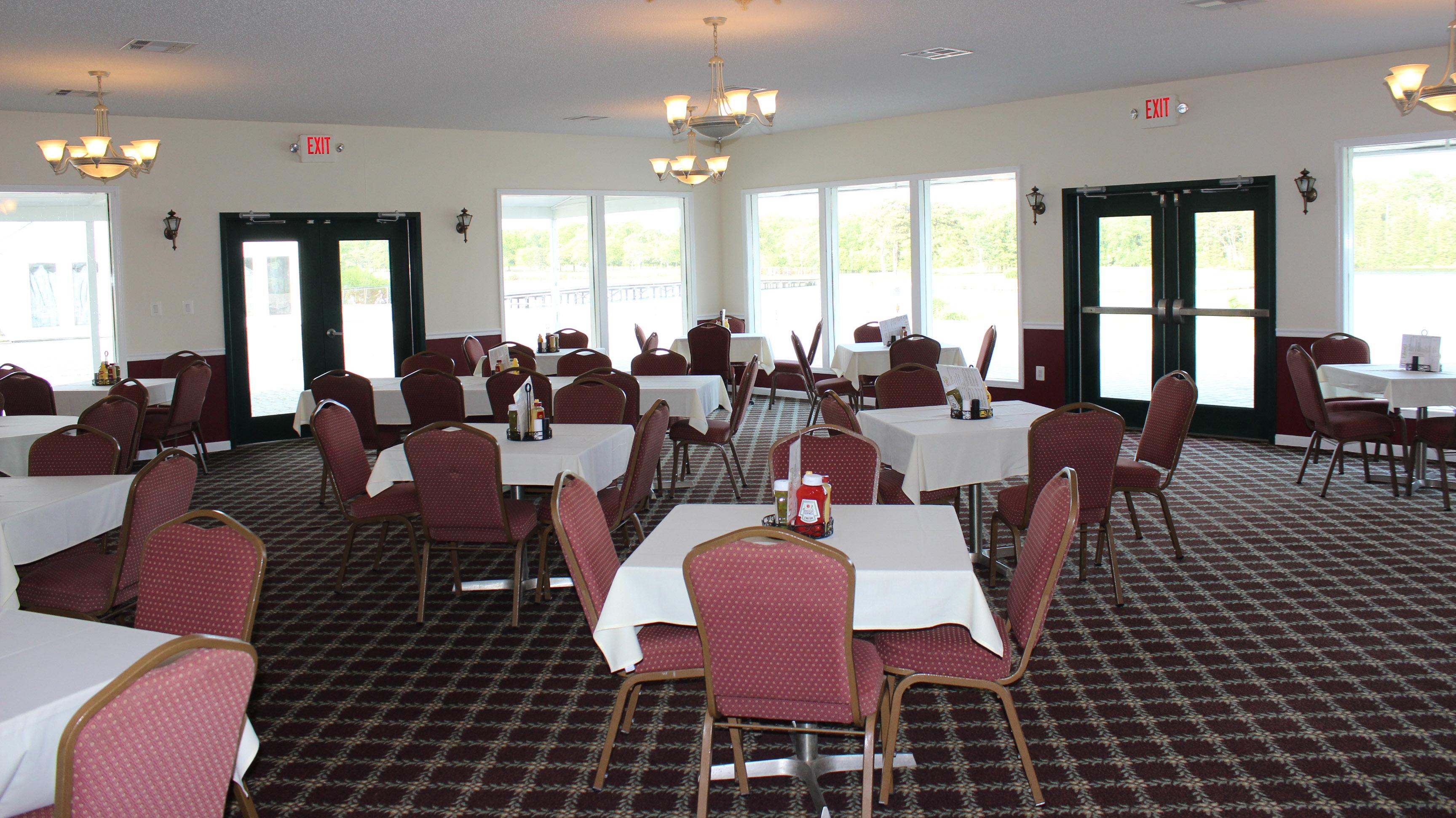 Several tables and chairs set with table cloths, condiments, and menus are arranged throughout the carpeted room. Various light fixtures and natural light from the large wall windows light the room. The tables are primarily four tops but there are also tables that seat groups of 6.