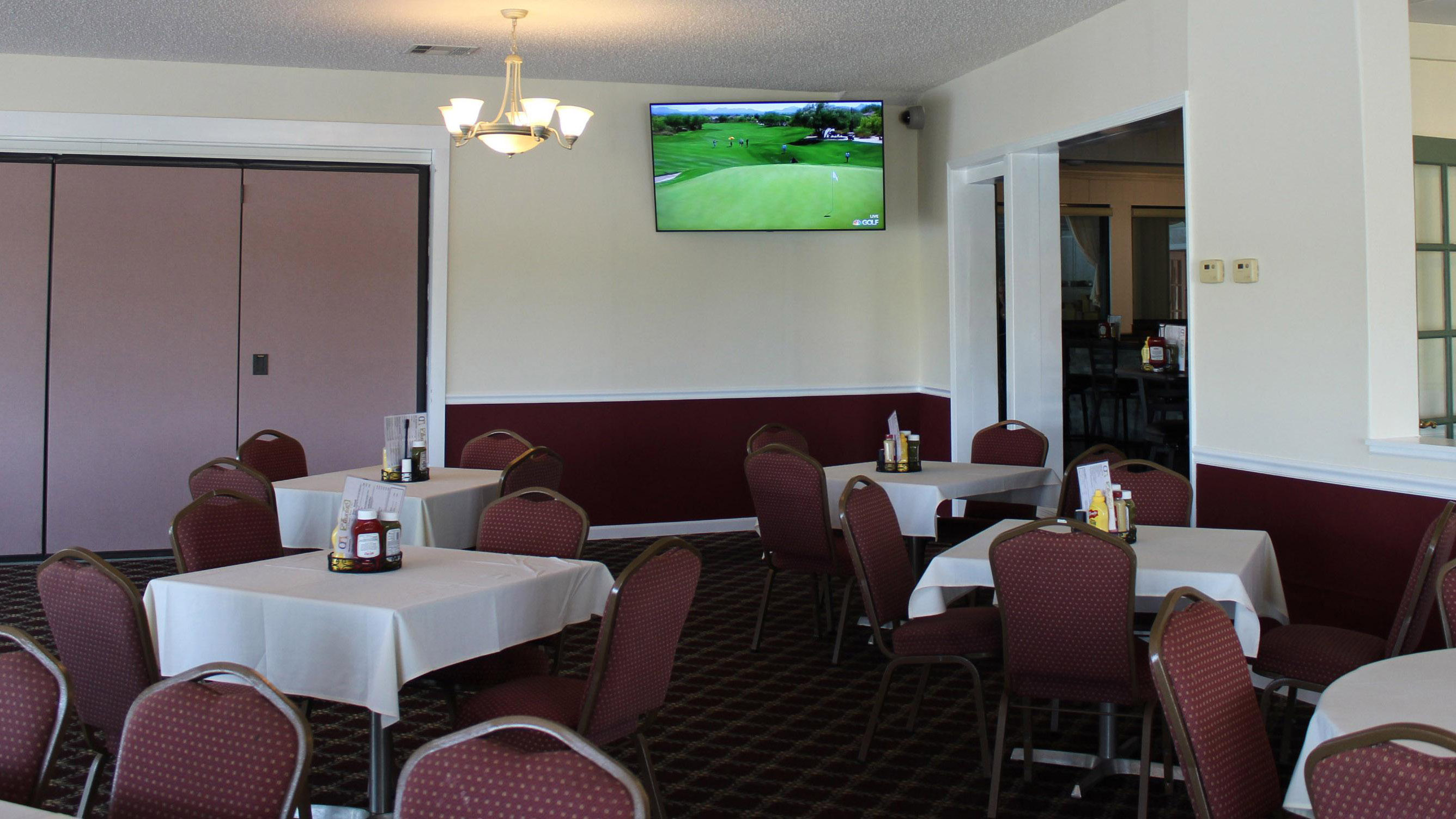 Dining room with several tables with table cloths and chairs set up. Condiments and menus are placed on the tables, and the television in the room is centered in the photo and is turned on to the Golf Channel.