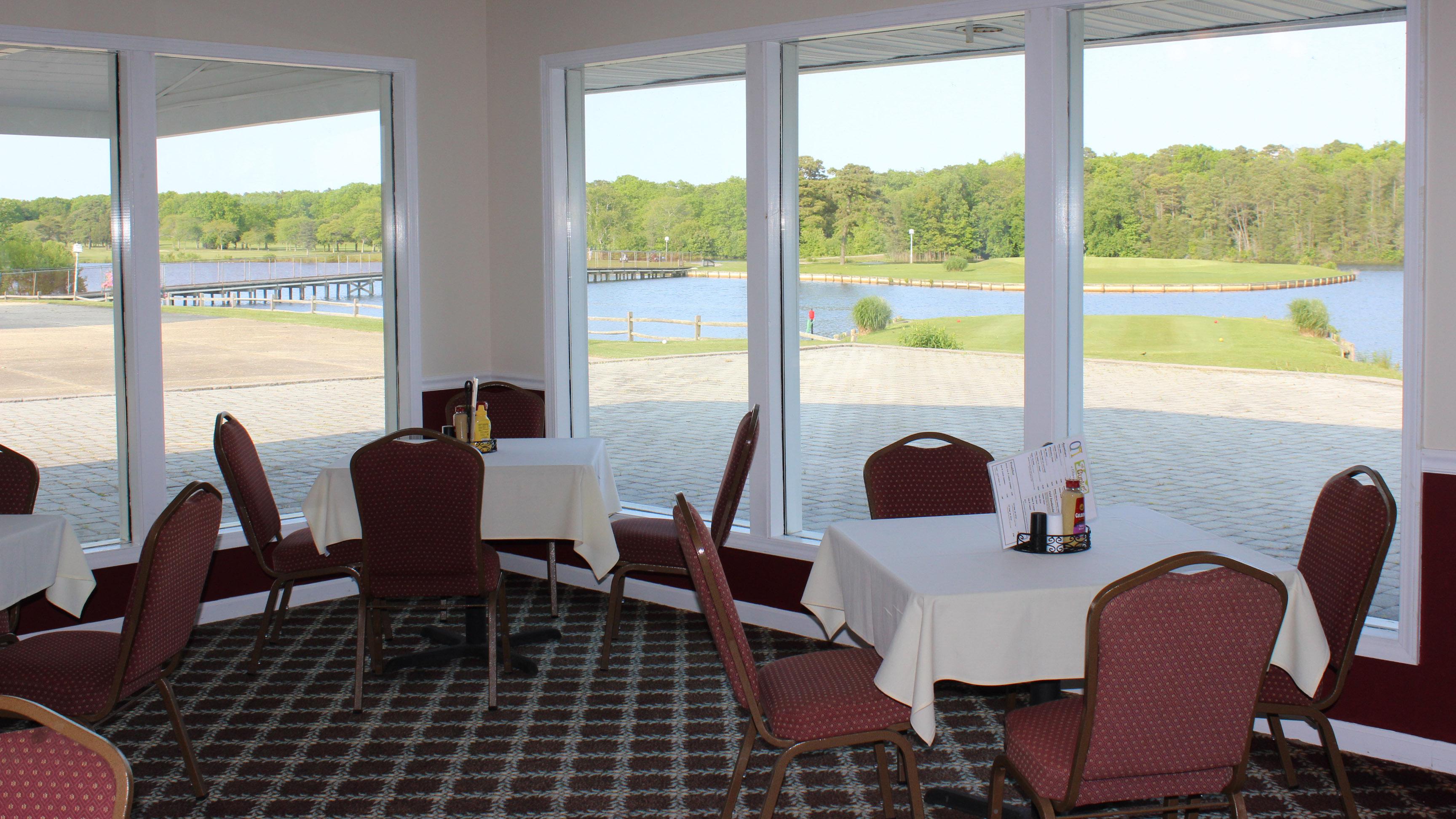 Two square tables with table cloths seating four with condiments and menus are positioned next to a series of wall windows. The golf course and lake can be seen through the windows.