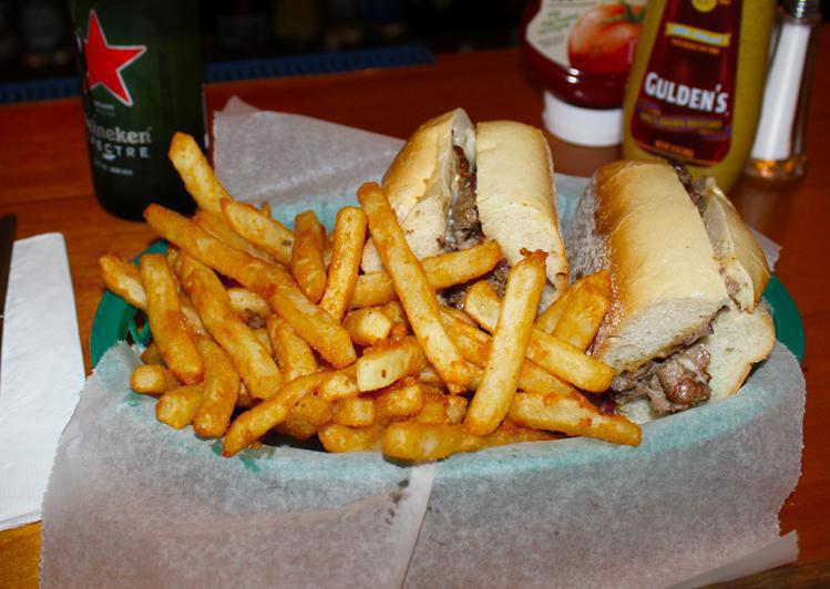 Cheesesteak in a steak roll cut in half and served with a good helping of french fries. A bottle of Heineken and Ketchup and Mustard bottles can be seen behind the food.