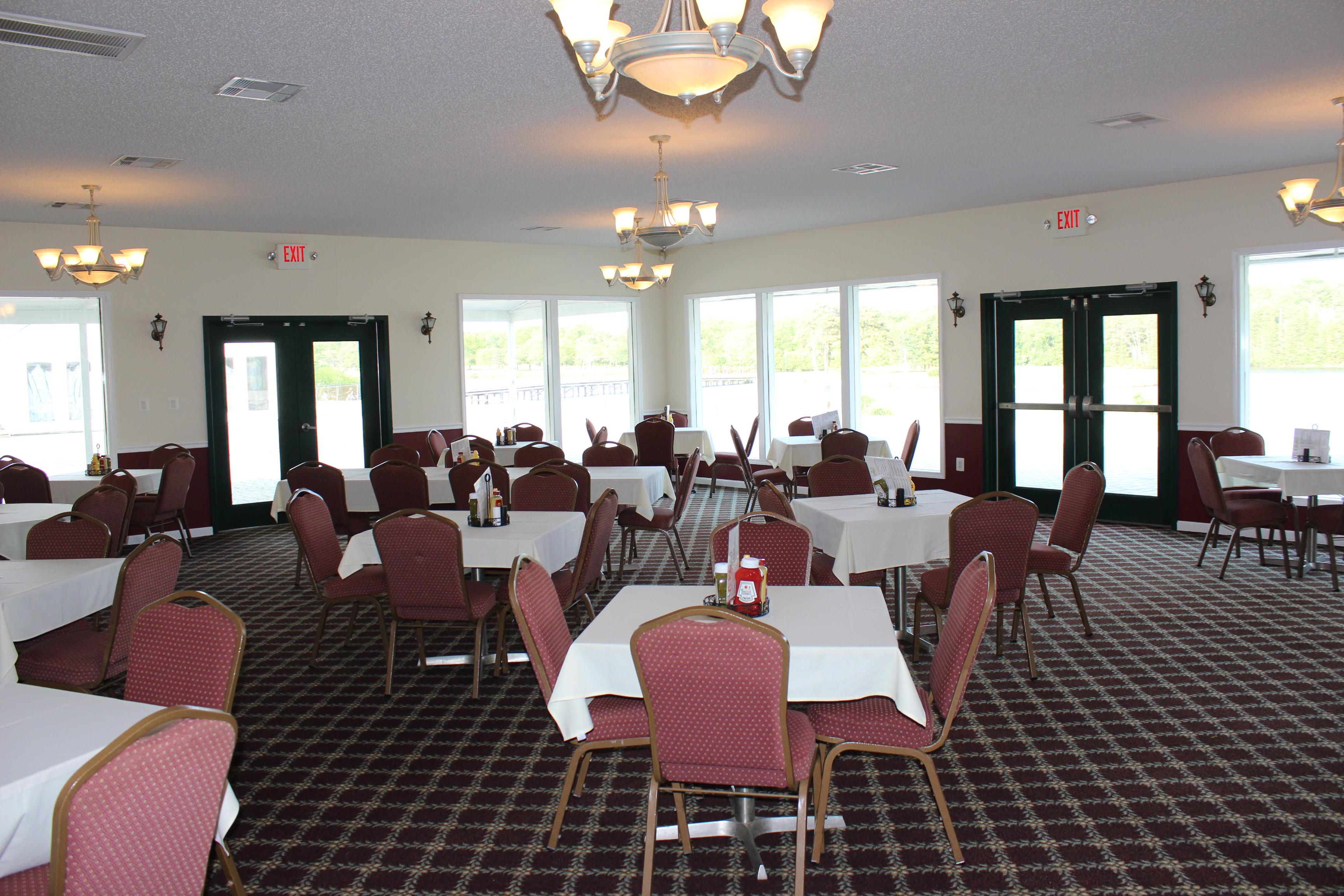Several tables and chairs set with table cloths, condiments, and menus are arranged throughout the carpeted room. Various light fixtures and natural light from the large wall windows light the room. The tables are primarily four tops but there are also tables that seat groups of 6.